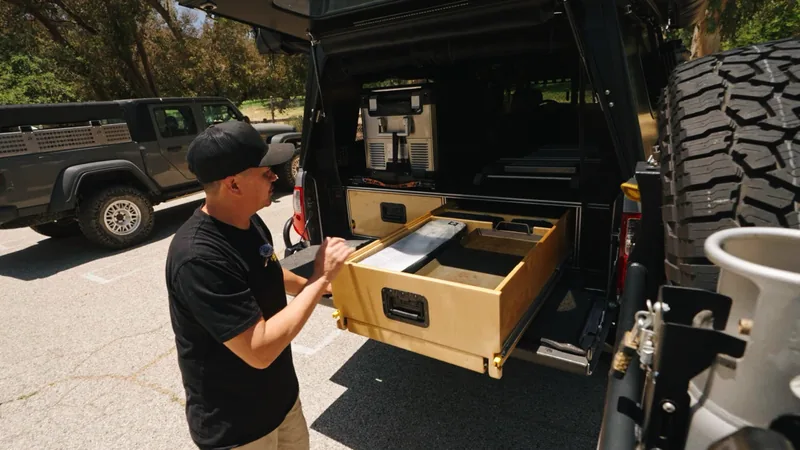 Man in a black cap pulling a large plywood storage drawer out from the truck bed camper, fridge visible behind it.