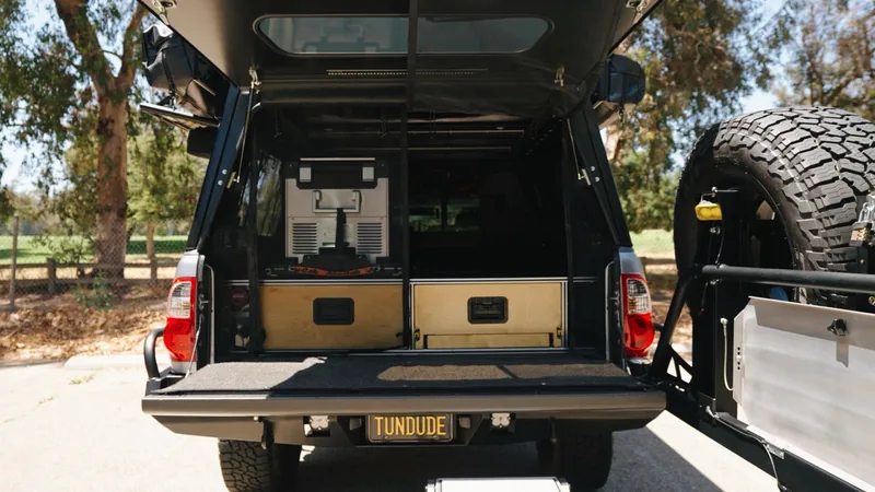 Rear view of the truck bed, hatch up and tailgate down, showing plywood drawers, a slide-out fridge and TUNDUDE plate.