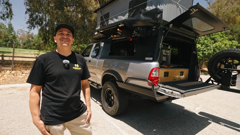 Smiling man in a black cap and tee standing in front of the pickup, camper roof raised and rear hatch open behind him.