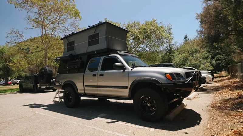 Silver Toyota Tundra pickup parked in a shaded lot with a grey pop-up wedge camper on the bed, roof raised.