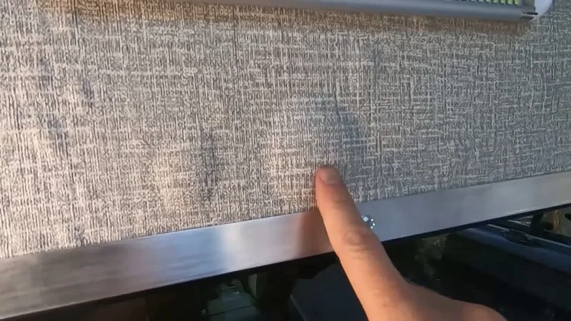 A finger points at a grey woven fabric panel just above a brushed metal rail inside the camper.