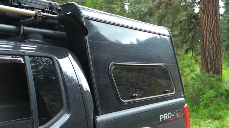 Close-up of the black camper shell's sliding side window and roof rack on a pickup parked among pine trees.