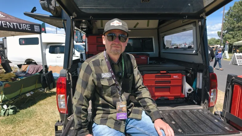 Man in plaid flannel and sunglasses sits on the truck's open tailgate, red tool chests stacked in the bed behind him.