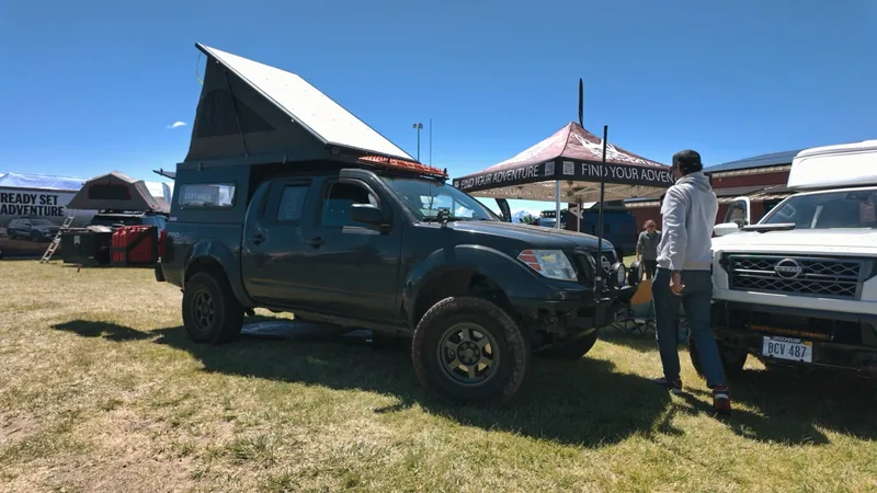Dark teal Nissan pickup on grass with its wedge camper roof popped up, at an outdoor expo with vendor tents.