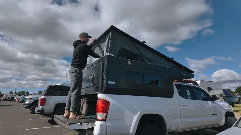 Man in a cap stands on the truck's open tailgate, reaching up to the side of the camper's raised pop-top roof.