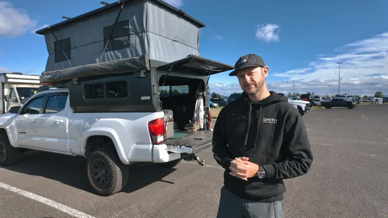 Man in a black Smith hoodie stands with hands clasped in front of the white pickup and its raised pop-up camper.